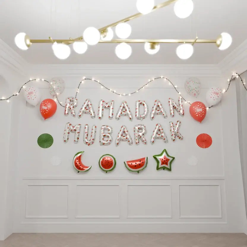 White watermelon-print Ramadan Mubarak foil balloons on a white wall with string lights and red balloons